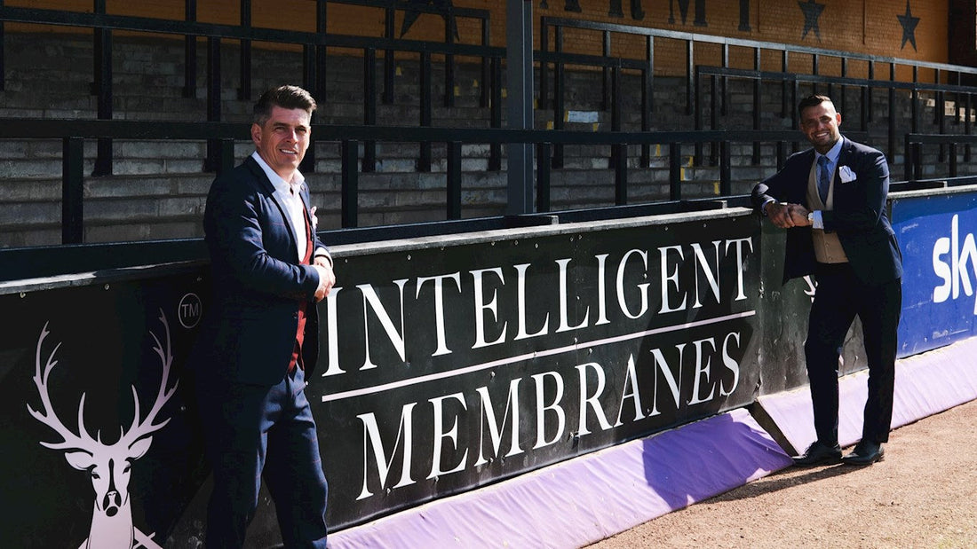 Adam White and club representative standing beside Intelligent Membranes sponsorship board at Abbey Stadium.