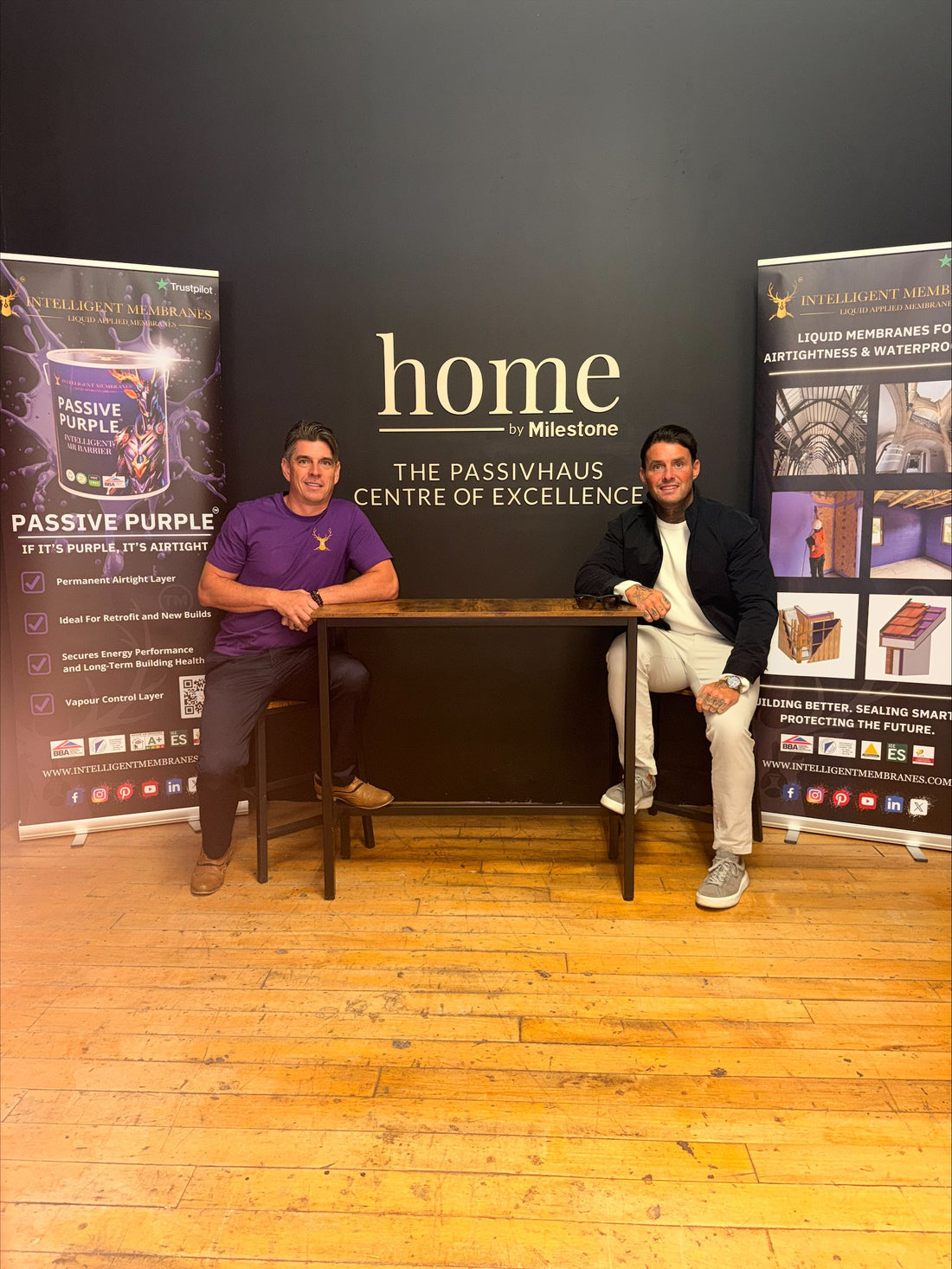 Two people seated beside Intelligent Membranes banners in front of the Home by Milestone sign.